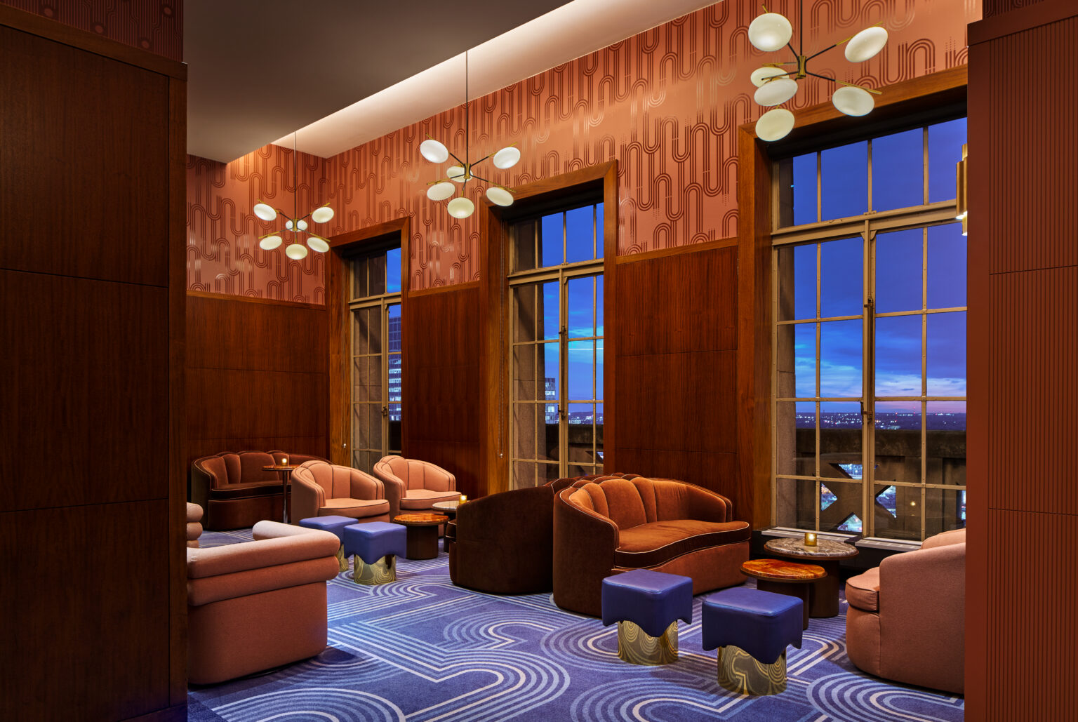 Modern lounge with blue-patterned carpet, brown sofas, orange walls, and large windows. Overhead, white, round chandeliers add elegance at dusk.