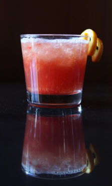 A glass of vibrant red cocktail with crushed ice, garnished with an orange twist, sits on a glossy surface, creating a dimly lit, sophisticated ambiance.