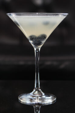 A martini glass filled with a light-colored cocktail garnished with blueberries. The background is dimly lit, creating an elegant ambiance.