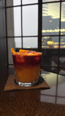A vibrant cocktail with layered orange and red hues, garnished with an orange peel, sits on a table by a window overlooking a cityscape at dusk.