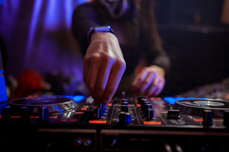A DJ adjusts the volume on a mixing console with a focused expression. The scene is lit in vibrant blues and purples, creating an energetic club atmosphere.