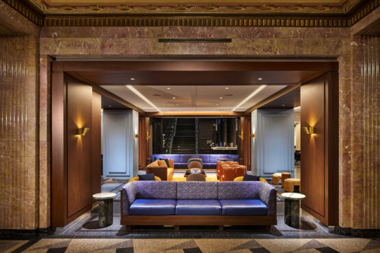 Elegant hotel lobby with a warm, inviting atmosphere. Features plush blue sofas, orange armchairs, and modern lighting against a marble and wood interior.