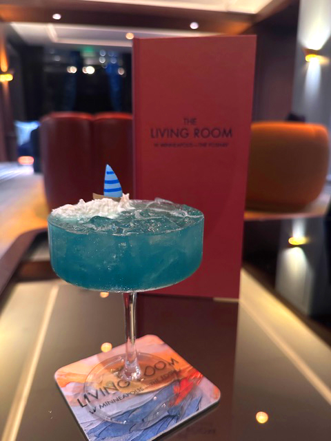 A vibrant blue cocktail in a glass, garnished with whipped cream and a small fin, sits on a branded coaster in an upscale lounge with soft lighting.