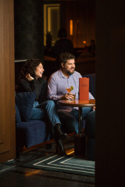 A couple sits in a dimly lit lounge, smiling and enjoying drinks. The man holds a cocktail, and a menu is on the table, creating a cozy atmosphere.