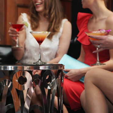 Women in elegant dresses enjoy colorful cocktails, seated around a stylish metallic table, laughing and chatting, conveying a joyful, social atmosphere.