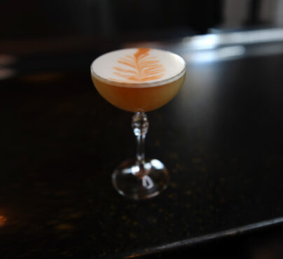 A frothy cocktail with a delicate leaf pattern sits in an elegant glass on a dark bar counter, evoking a sophisticated and inviting atmosphere.