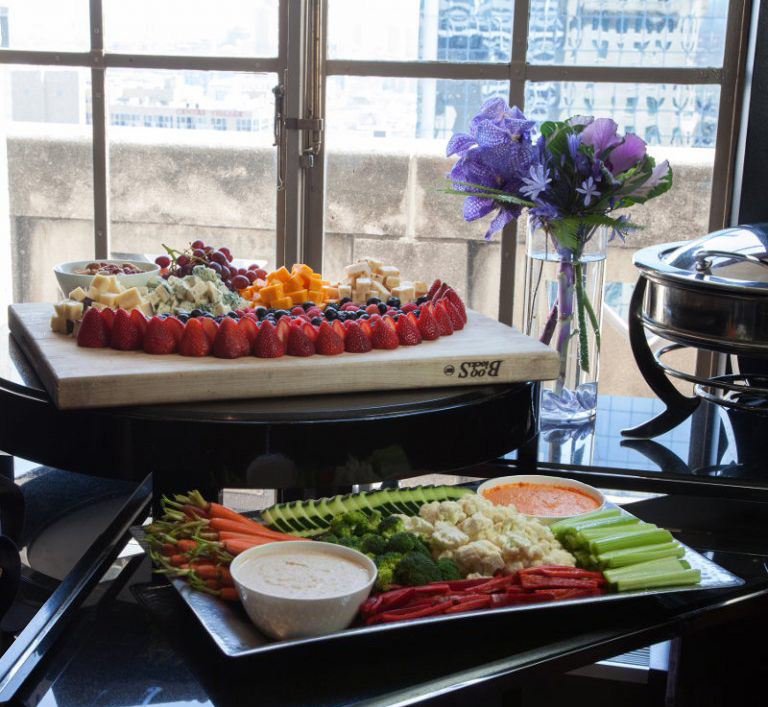 A vibrant spread of fruits and vegetables on a table by a large window. A wooden board displays assorted fruits and cheese, with flowers nearby.
