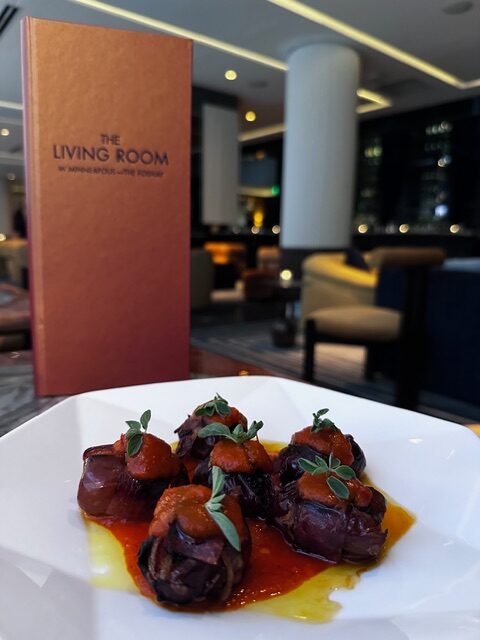 Plate of gourmet appetizers topped with herbs on a white dish, set against an elegant restaurant backdrop with soft lighting and a "The Living Room" menu.