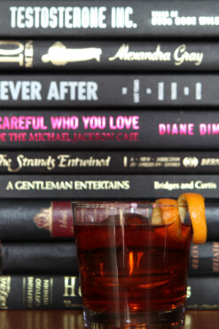 A stack of books with varied titles in a warm tone backdrop, centered behind a glass of amber-colored cocktail garnished with an orange twist.